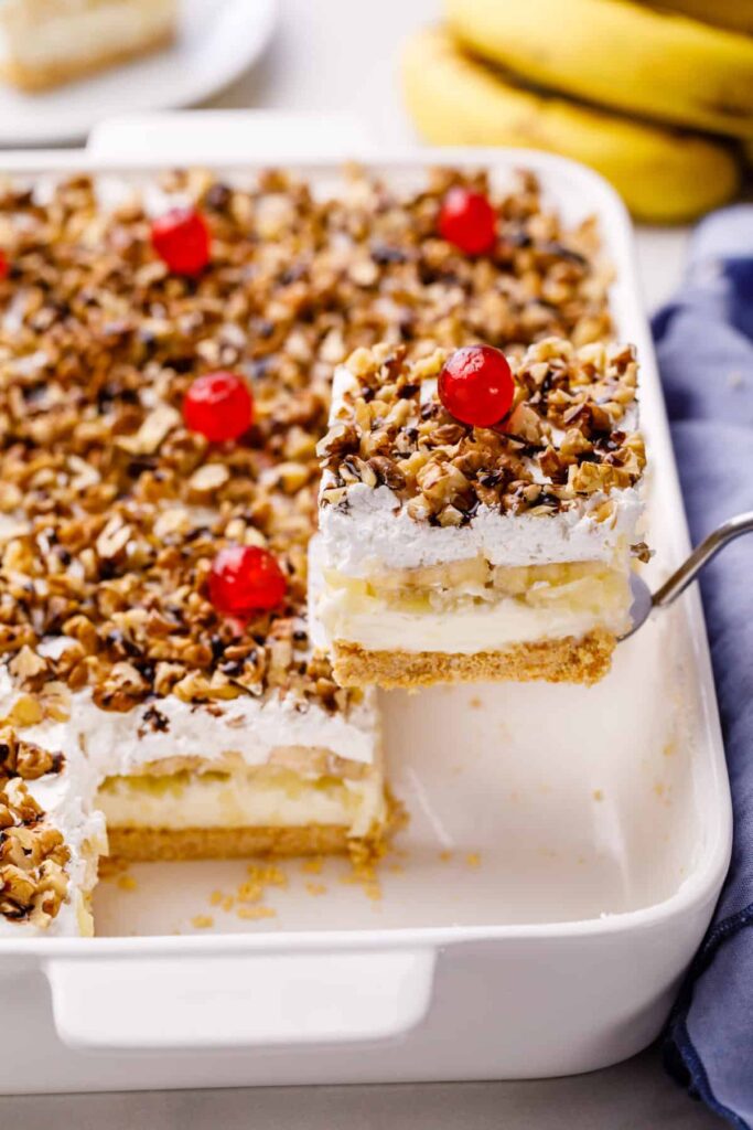A creamy, layered no-bake banana split cake topped with fresh banana slices, crushed pineapple, whipped cream, cherries, and chopped nuts.