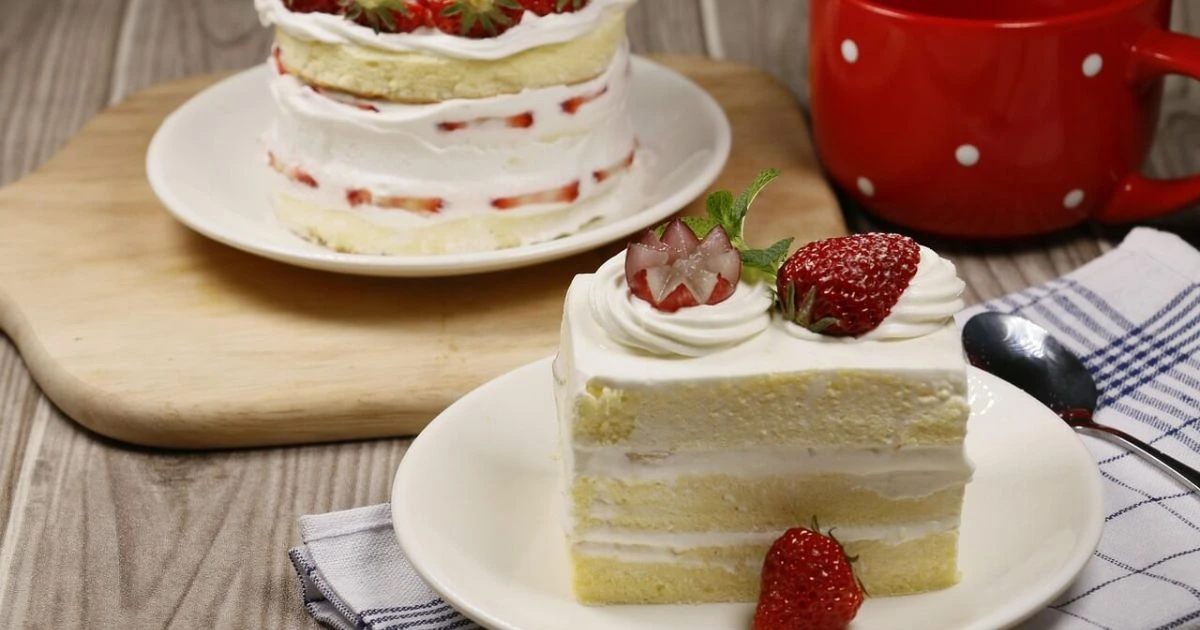 Slice of layered strawberry shortcake with white frosting and fresh strawberry garnish alongside a whole cake on wooden serving board and red polka dot mug"