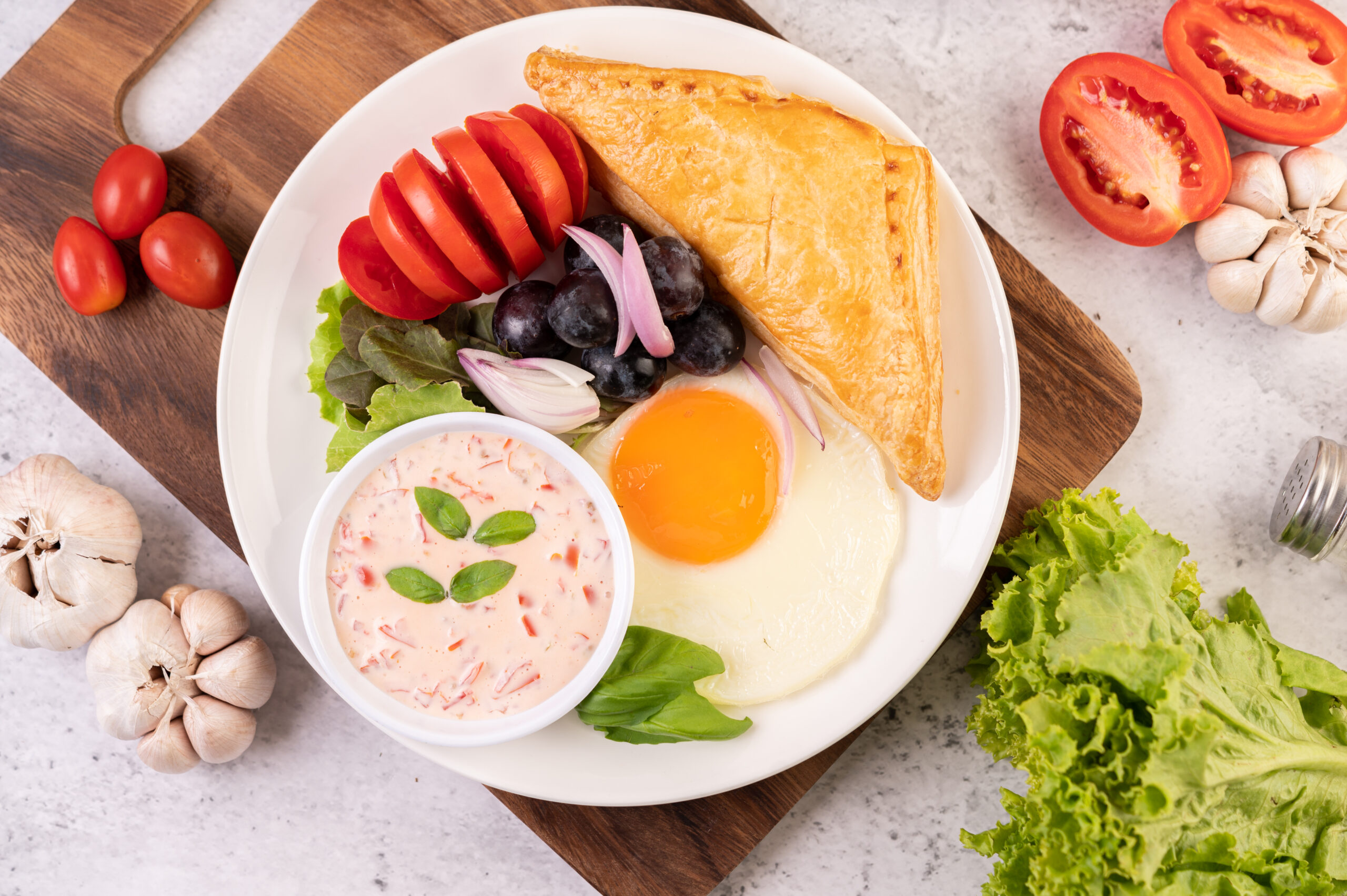 Breakfast consists of bread, fried egg, salad dressing, black grapes, tomatoes, and sliced ​​onions.