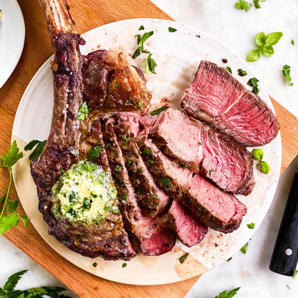 Grilled tomahawk ribeye steak sliced on a wooden cutting board, served with roasted asparagus, lemon wedges, and fresh rosemary.