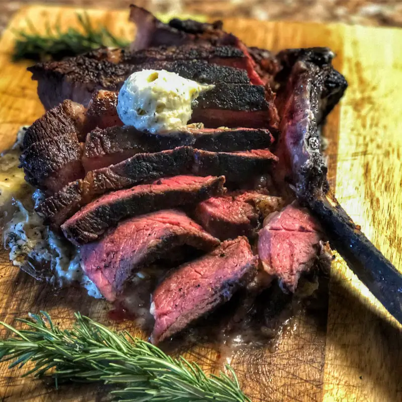 Raw tomahawk ribeye steak on a dark background, seasoned with coarse salt, cracked pepper, and garnished with fresh herbs.